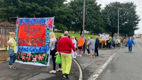 Demonstrators on a pavement outside a Heol Goffa school. There are about 30 people in the pictures, some holding a banner saying protect care love Ysgol Heol Goffa