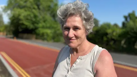 Tom Jackson/BBC Head and shoulders image of Juliette Jackson in front of the red road on Adams Road. She has short grey hair and is wearing a white sleeveless top.