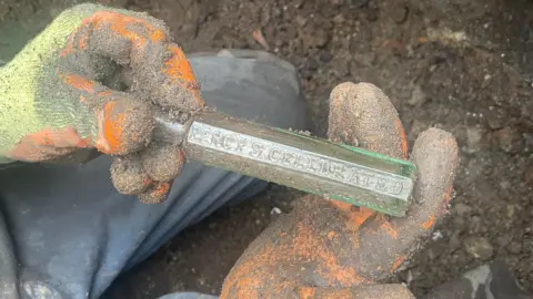 Andy Matthews Gloved hands holding a small, rectangular glass bottle freshly dug from dark soil. The bottle is embossed with the words 'Percy's Celebrated'.