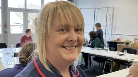 Saoirse Read is smiling and has blonde hair. She's in a room with other people and a bank of tables and chairs. She is in a nursing uniform. 