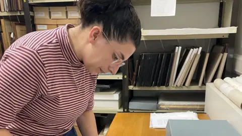 Dacorum Heritage Trust Connie Search, wearing a red and cream striped top, a necklace and glasses and she has her hair tied up. Files are behind her on shelves, and she is looking down at a table top, with items on it. 