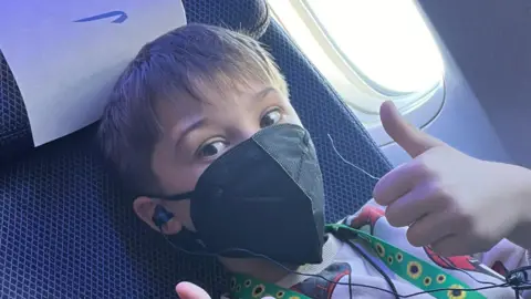 Katie Hutt Yann Jennings, a 10-year-old boy, sat in an airplane seat wearing a black face mask, headphones, and a green lanyard with sunflowers on it. Giving thumbs up to the camera. 