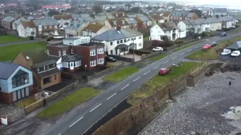A aerial shot of the seafront road at Beadnell. Large houses are on one side of the road, facing a pebbly beach on the other. Behind are a few hundred other houses with the sea and a stretch of the coastline in the top far right. 