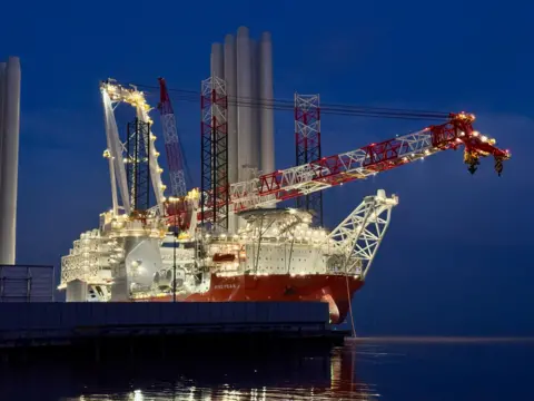 An image of the Wind Peak taken at night. The dark skies are illuminated by lights on the red and white vessel which has a long crane spanning the length of the deck.