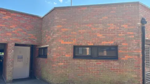 Brick toilet block with a white door, with a sign on it, windows and a down pipe.