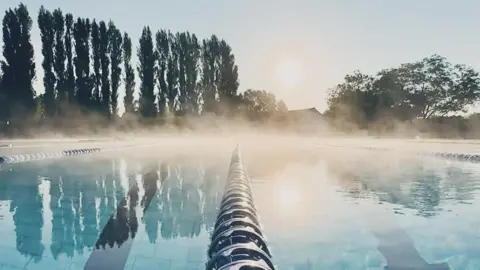 The pool, with mist above its surface, a line of fir trees in the distance, and the sun rising on the horizon.