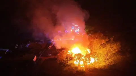 Dan Jessup Aerial night-time image showing a large fire engulfing farm buildings, with bright flames and thick smoke rising into the air as emergency vehicle lights illuminate the surrounding countryside.