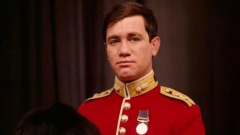 National Portrait Gallery A colour picture showing Army officer Captain Robert Nairac, He has short, brown hair and is wearing a red military jacket decorated with gold buttons, gold epaulettes and single medal pinned to his chest. He is holding a tall, black bearskin hat.
