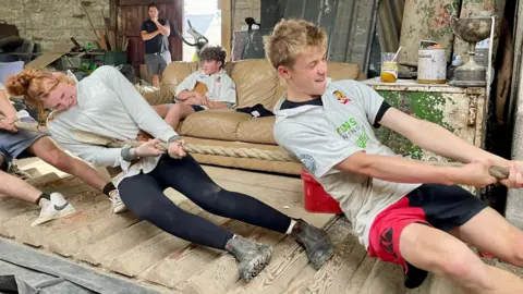 BBC A teenage boy and girl pull on a tug of war rope. The boy has short blonde hair, wearing a white polo with various printed logos and a pair of bred and black shorts. The girl has curly red hair tied in a bun, a white rugby shirt, black leggings and muddy black boots. They are standing on a heavily grooved surface inside an untidy barn. A boy with curly hair and a white polo is sat on a cream settee in the background. A man in a black t-shirt stand behind him watching the pullers.