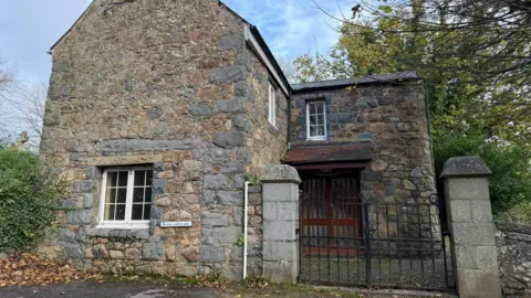 BBC "The Barn" next to St Andrew's Church. The photograph is taken from outside the property. There is a black gate to the property's front door, which looks like vanished wood. The exterior of the property is old brick work with white sash windows.