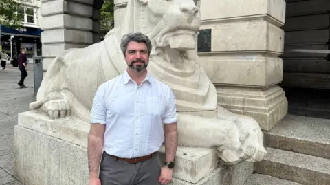 Nottingham City Council Man stood in front of stone lion statue. He is wearing a white shirt and smiling.