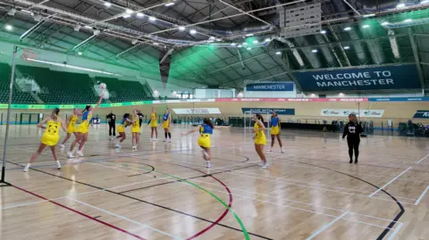 Manchester Thunder players on the netball court wearing yellow sports dresses. Half of them wear blue bibs. One player is jumping to block the ball.