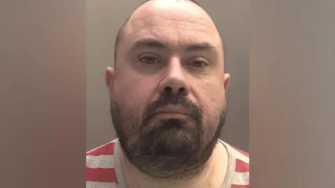 Police mugshot of Peter Stanley wearing a red and grey striped T-shirt. He has a balding head and a black beard. 