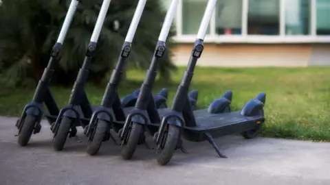 A row of five black scooters parked on a pavement next to a grassy area