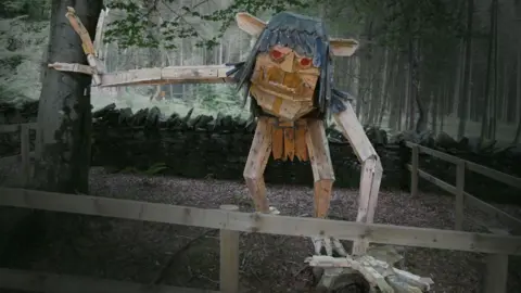 CULTURE VANNIN A wooden sculpture of the Buggane, which has long dark hair with its ear sticking out, red eyes and protruding teeth, along with long arms.