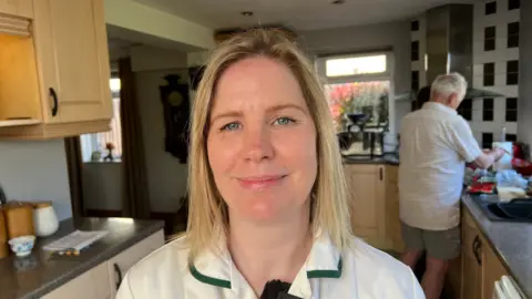 Woman with blonde hair smiling softly while a man potters in the kitchen behind her. The woman is wearing a white uniform.
