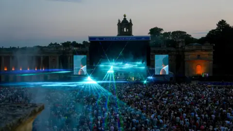 Blenheim Palace Festival A stage with lights coming off it sits in front of Blenheim Palace. There is a large crowd watching on. It is twilight.