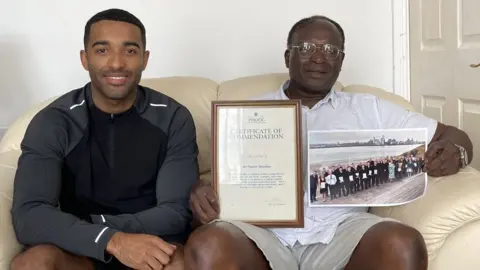 BBC Nelson Shardey sits on his sofa holding his police commendation certificate, alongside his son Jacob