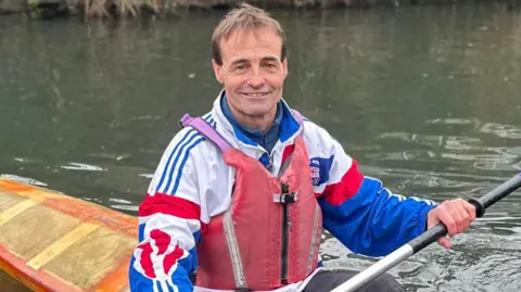 A Caucasian man sitting inside an orange kayak boat holding a black and blue paddle stick. The boat is in the canal. The man is wearing a Team GB jacket that is white, red and blue. He is also wearing a red life jacket. 
