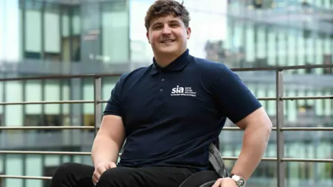 Spinal Injuries Association Dave Eastham is smiling at the camera whilst sitting in his wheelchair. He has brown hair and is wearing a t-shirt with the Spinal Injuries Association logo