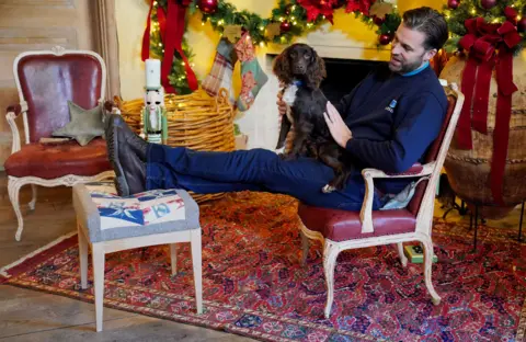 PA Media A dog from Battersea Dogs and Cats Home with a male handler who has his feet up on a stool covered in curtains recycled from Buckingham Palace