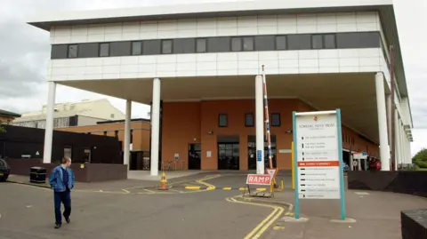 The entrance to a hospital building, with a sign up displaying directions for where to go for different departments. A man wearing a denim jacket is walking by, looking back at the hospital. 
