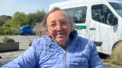 An old woman with grey hair and glasses smiles at the camera, wearing a light blue puffer jacket. Behind her is a white van, a blue car and greenery.