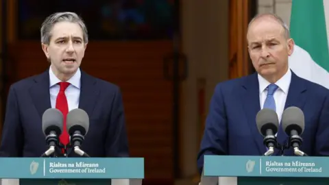 Simon Harris and Micheál Martin both stand in front of green Government of Ireland branded podiums. Mr Harris is wearing a navy suit, light blue shirt and red tie. Mr Martin is wearing a dark blue suit, white shirt and dark blue tie.