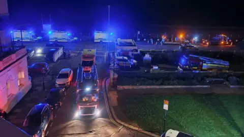 A street view of a residential area. There are emergency services and cars parked near some flats during the night.