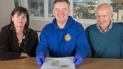 MNH Manx National Heritage Curator Allison Fox, who has shoulder-length dark brown hair and is wearing a black jacket, sitting at a table next to finder Rob Middleton, who has short grey hair, is wearing a blue hoodie and blue latex gloves and holding the spoon on a piece of paper, and landowner David Anderson, who is wearing a blue jumper and shirt and has short grey hair.