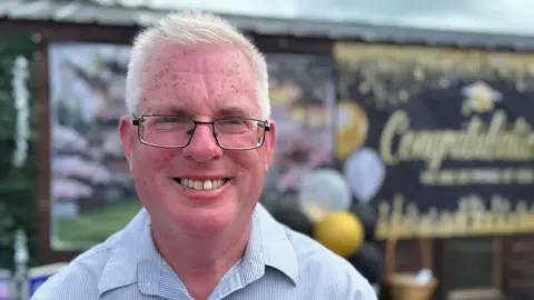 An older man with short white hair He's rectangular glasses with a thin black frame and is smiling. Behind him is a very large banner which reads 'Congratulations'. There are also yellow, black and white balloons along with other decor.