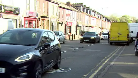 BBC A street scene showing a row of terraced houses, several of which have shops built into the bottom floor. There are cars driving down the road and some parked on the pavement