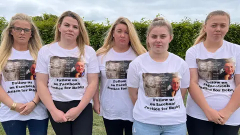 Five sisters, side-by-side. There are all looking to the camera. They are all wearing jeans and have the same white T-shirt on. It states "Follow us on Facebook. Justice for Bobby". They are standing on grass and there is a green hedge behind them. 