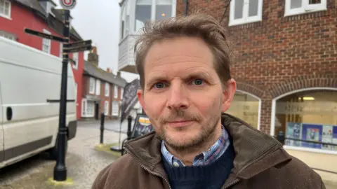 Shows a man with short brown hair and short beard. He is standing outside on the street with a brick building behind him. He is wearing a brown jacket over a blue jumper.