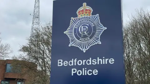 A close up shot of the large blue sign outside Bedfordshire Police headquarters. It has the police crest on and the words Bedfordshire Police in white font. There is a red brick building to the left, and bare trees behind.