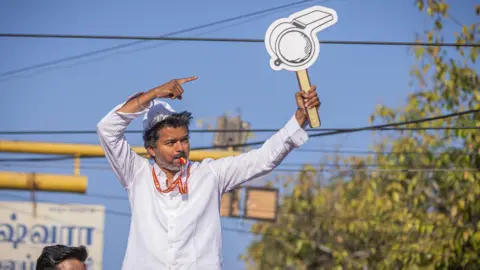 TVK Vijay, wearing a white shirt with a whistle in his mouth, poses with his party's symbol
