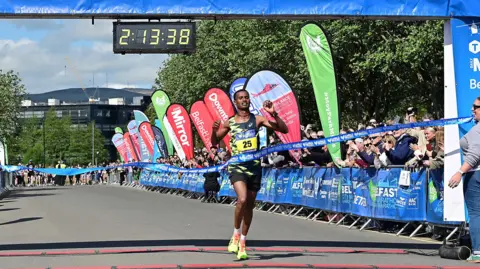 Pacemaker Press A man with short black hair crossing the finish line. He is wearing a black and yellow running vest, black shorts and yellow trainers. He is crossing through a blue ribbon and you can see the crowd cheering him on from behind a blue branded baricade.
