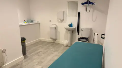 Supplied A large disabled toilet containing a toilet, a hoist, a changing bench and a baby changing table. 