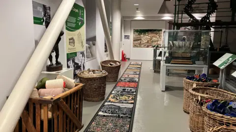 A museum, exhibition, with artwork and photographs of carpets and buildings on the floor and baskets containing yarn. There are information plaques on the walls and a display cabinet in the distance.