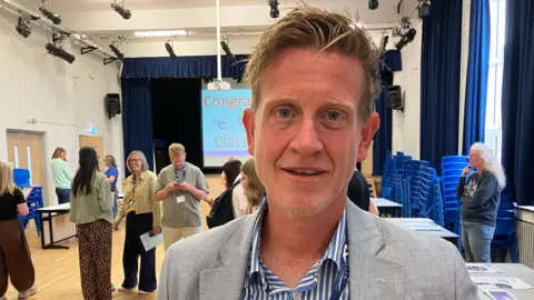 Seb Cheer/BBC A man in a school hall wearing a light grey blazer and a blue‑and‑white striped shirt stands in the foreground with a staff lanyard, while groups of people talk near tables.
