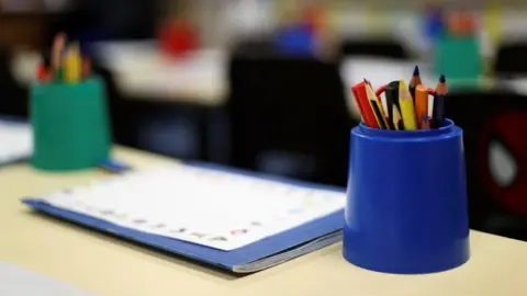Pencils are pictured in a blue pot on a desk in an empty classroom. To the left is a blue school exercise book with a piece of paper on the top and a green pencil pot in the background.