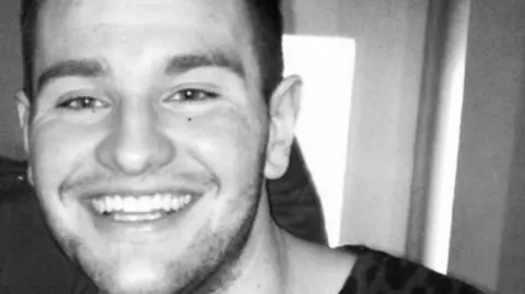 Family Handout The image shows a black‑and‑white photo of the upper body and face of Daniel Baird. Only part of his head, ear, neck, and shoulder are visible. Daniel is wearing a patterned top, and the background includes a door with a small sign above it.