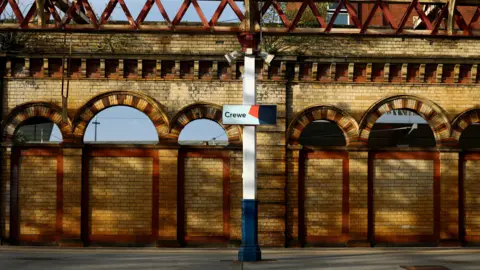 Reuters A railway platform with a brick wall of brown and orange bricks at the side of the platform, including a series of arches which are open to show the outside world of a blue sky. A metal post stands in the centre of the platform, painted white with a blue stump at the bottom. On the post is a sign with "Crewe" written on it.