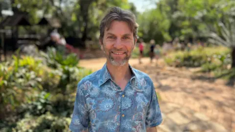 Marc is a white man with brown and grey hair, and a small beard. He wears a brightly coloured blue shirt and smiles into the camera. The picture is taken outside, and he is surrounded by greenery.