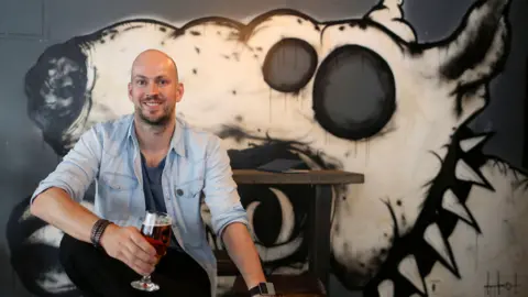 Getty Images James Watt pictured in 2015. He is holding a beer and sitting in front of a wall which is painted with a picture of a white dog wearing a studded collar.