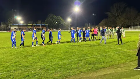 Contributed Two sets of football players are shaking hands on the pitch. One team is wearing blue and white, the other team has green and white striped tops. 