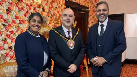 City of Wolverhampton Council A woman and man stood either side of the mayor who is wearing a mayoral chain, suit and tie. The woman, who is on the left, is smiling and wearing a blue jacket. The man, who is on the right of the mayor, is also smiling and is wearing a blue suit and tie. There is flowery wallpaper in the background. 