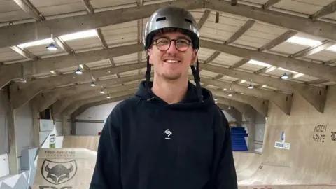 James Burridge/BBC Jayden Sharman smiling into the camera at Adrenaline Alley skatepark. He has a grey helmet on, round framed glasses and a black hoodie. Large wooden ramps are visible in the background.