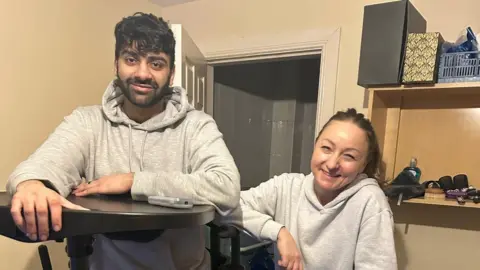 Yousaf Ghaffar Two people stand, smiling at the camera. The man on the left has dark hair and facial hair. He is wearing a grey hoodie and is leaning on a black contraption. The woman on the right has dark hair and is also wearing a grey hoodie. 