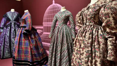 Four period dresses are on display. The walls behind are maroon